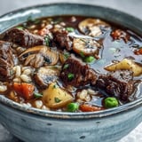 A pot of beef and barley soup simmers on the stove, filled with mushrooms, potatoes, and fresh herbs.