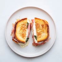 Golden sourdough grilled cheese with crispy prosciutto and creamy burrata, sprinkled with fresh basil on a rustic plate.  